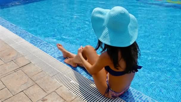 Mladá žena v plavání oblek a modrý klobouk sedí na kraji bazénu a opalovat odběr - Záběry, video