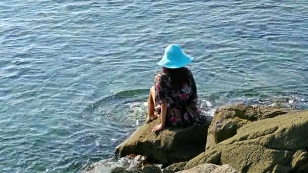 Mujer joven en vestido de verano y sombrero azul sentado en las rocas en la orilla del mar
 - Metraje, vídeo