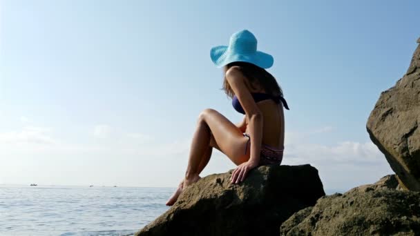 Mujer joven en traje siwmming y sombrero azul sentado en las rocas en la orilla del mar
 - Metraje, vídeo