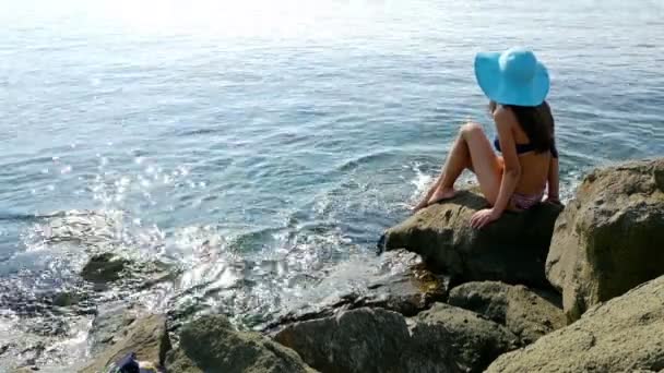 Mujer joven en traje de baño y sombrero azul sentado en las rocas en la orilla del mar
 - Metraje, vídeo