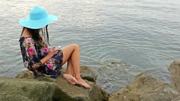 Mujer joven en un vestido de verano y sombrero azul sentado en la costa rocosa del mar y tomar una foto de sí misma con el teléfono celular
 - Metraje, vídeo