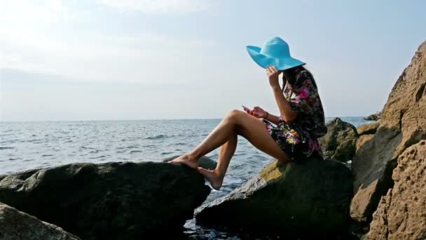 Mujer joven en un vestido de verano y sombrero azul sentado en la costa rocosa del mar, teléfono inteligente en sus manos
 - Metraje, vídeo