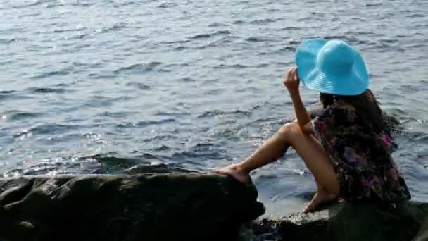 Silueta de una joven con un colorido vestido de verano y sombrero azul en la costa rocosa del mar
 - Metraje, vídeo