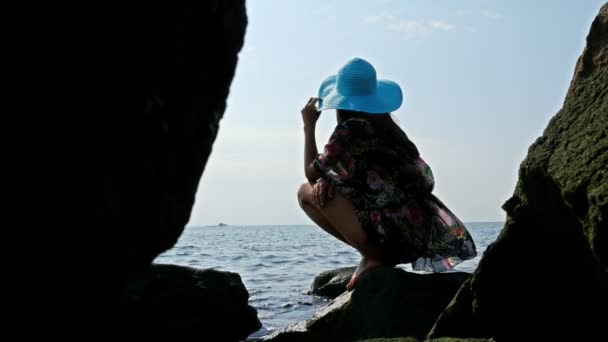 Silueta de una joven con un colorido vestido de verano y sombrero azul en la costa rocosa del mar
 - Metraje, vídeo