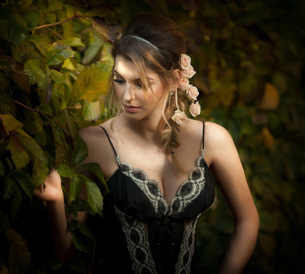 Beautiful sensual woman with roses in hair posing near a wall of green leaves. Young female in black elegant dress daydreaming in nature. Attractive voluptuous lady with creative hair arrangement - Photo, Image