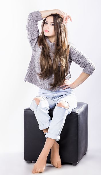 belle fille en pull tricoté assise sur une perruque noire sur un fond blanc. Cheveux longs et foncés. jeans déchirés
 - Photo, image