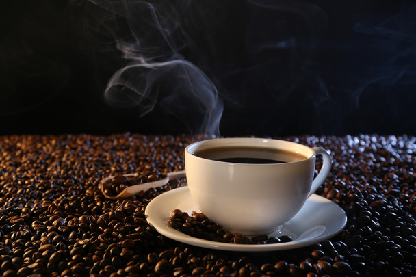 Kop warme koffie onder koffie bonen op donkere achtergrond - Foto, afbeelding