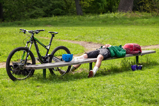 Велосипедист відпочиває на лавці в парку. Втомився молодий чоловік зі своїм гірським велосипедом у лісі відпочиває і робить паузу
. - Фото, зображення