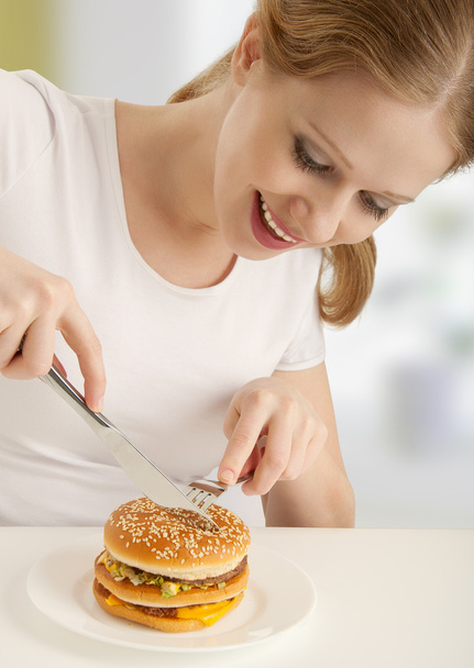 ελκυστικό κορίτσι με ένα πιρούνι και μαχαίρι τρώει χάμπουργκερ - Φωτογραφία, εικόνα
