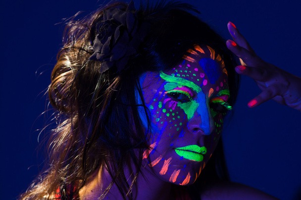 Headshot woman wearing awesome glow in dark facial paint, blue based with other neon colors and obscure abstract background, facing camera - Zdjęcie, obraz