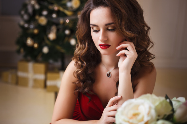 Close-up portrait of beautiful young woman in gorgeous red velvet evening dress sitting by the table in expensive interior - Fotografie, Obrázek