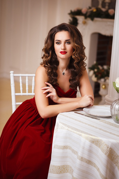 Close-up portrait of beautiful young woman in gorgeous red velvet evening dress sitting by the table in expensive interior - Foto, Imagen