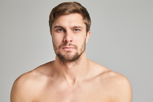 Portrait of a serious young man with bare shoulders on a gray background, powerful swimmers shoulders, beard, charismatic, adult, brutal, athletic - Valokuva, kuva