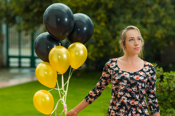 schönes hispanisches Model trägt Sommerkleid in Gartenumgebung und hält goldene und schwarze Luftballons hoch, während sie für die Kamera posiert - Foto, Bild