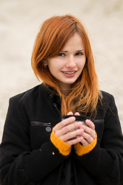 Beautiful red-haired girl in a black fashionable coat  trying to keep warm in the cold weather.Cheerful smiling red-haired girl with cute smile with a cup of hot tea, coffee, chocolate.Cheerful smiling red-haired girl with cute smile red hot coffee. - Photo, Image