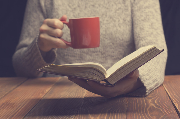 Young woman reading a book and holding cup of tea or coffee. Toned image - Photo, Image