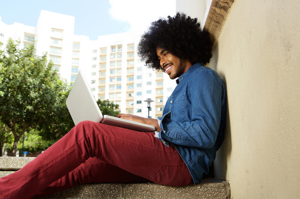 Hymyilevä miesopiskelija istuu ulkona työskentelemässä kannettavan tietokoneen kanssa
 - Valokuva, kuva