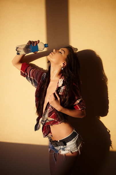 Retrato de moda bonita menina morena sexy em jeans e camisa colorida por trás do pôr do sol com água de garrafa dentro de casa
 - Foto, Imagem
