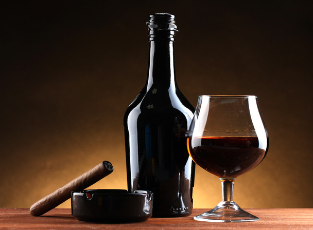 Bottle and glass of brandy and cigar on wooden table on brown background - Photo, Image