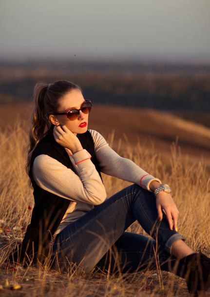 Приваблива, приваблива, серйозна дівчина з сонцезахисними окулярами, що сидить на пагорбах з красивими видами дивитися захід сонця
. - Фото, зображення