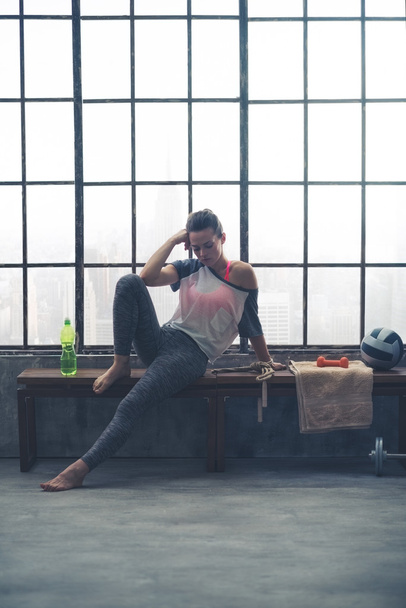Pensive woman in workout gear resting on bench in loft gym - Фото, зображення