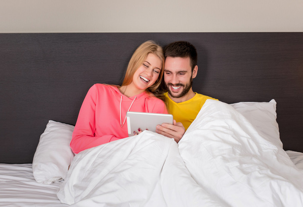 Молодая сладкая пара в постели Смотря что-то на планшетном гаджете
 - Фото, изображение
