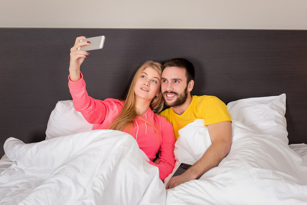 Nuori onnellinen pari sängyssä tekee selfie puhelinkameralla
. - Valokuva, kuva