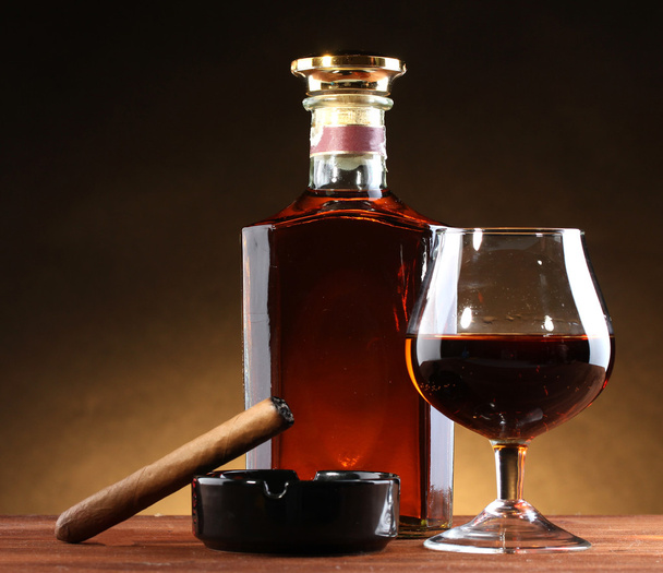 Bottle and glass of brandy and cigar on wooden table on brown background - Fotoğraf, Görsel