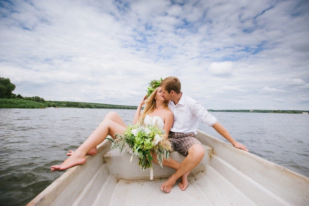 Happy young couple with a bouquet and a wreath hug sitting in a boat on the lake - Fotografie, Obrázek