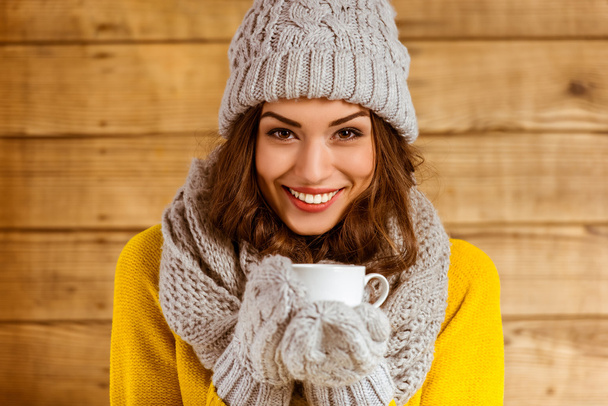 Portrait of a beautiful young girl with cap and gloves drinking tea on the background of wooden wall - Foto, Imagen