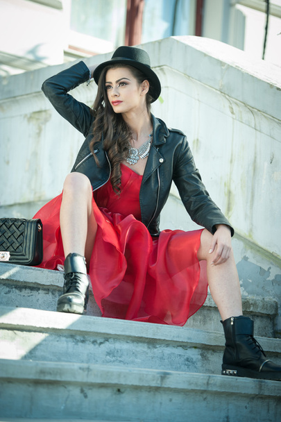 Beautiful woman with black hat, red dress and boots posing sitting on stairs. Young brunette spending time during autumn. Long hair attractive girl with creative makeup and red dress, outdoors shot - Φωτογραφία, εικόνα
