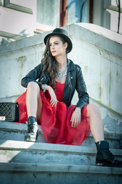 Beautiful woman with black hat, red dress and boots posing sitting on stairs. Young brunette spending time during autumn. Long hair attractive girl with creative makeup and red dress, outdoors shot - 写真・画像