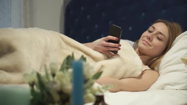 Ξανθιά ξαπλωμένη στο κρεβάτι, πληκτρολογώντας ένα μήνυμα στο τηλέφωνό σας - Πλάνα, βίντεο