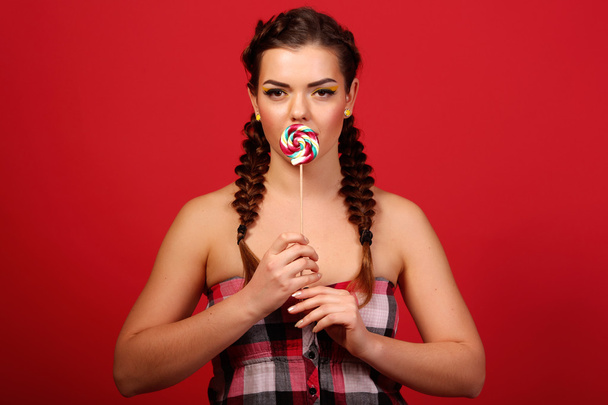 Beauty fashion model girl eating colorful lollipop. Surprised funny young woman with braids hairstyle, nails and makeup beige, isolated on a red background - Photo, Image
