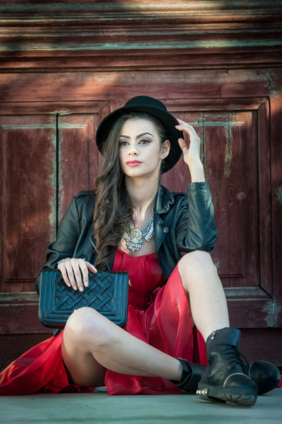 Young beautiful brunette woman with red short dress and black hat posing sensual in vintage scenery. Romantic mysterious young lady relaxing sitting on floor against vintage wooden wall - 写真・画像