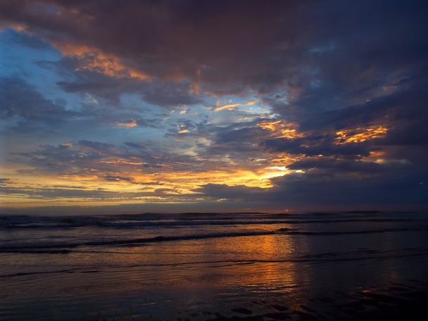 Pôr-do-sol nublado sobre o oceano com ondas em primeiro plano
 - Foto, Imagem