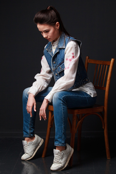 грустная брюнетка в джинсовом жилете сидит на стуле
 - Фото, изображение