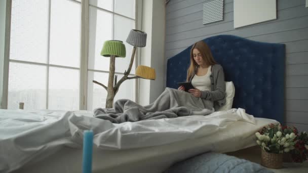 Młoda dziewczyna siedzi na łóżku z tabletem - Materiał filmowy, wideo