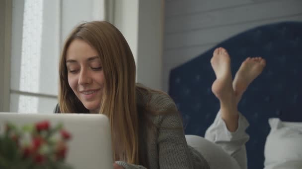 Χαμογελαστό κορίτσι ξαπλωμένο στο κρεβάτι με έναν φορητό υπολογιστή - Πλάνα, βίντεο