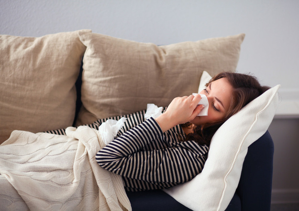 Portrait of a sick woman blowing her nose while sitting on the sofa - Photo, Image