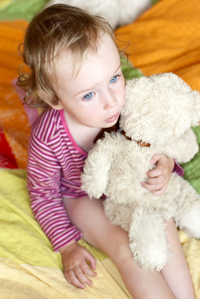 Petite fille triste aux yeux bleus assise sur un lit avec un ours en peluche
. - Photo, image