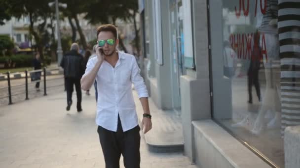 . Młody mężczyzna ubrany w okulary przeciwsłoneczne i spacerów po centrum miasta i rozmawiając telefonicznie. Miejski mężczyzna w jego 20s. - Materiał filmowy, wideo