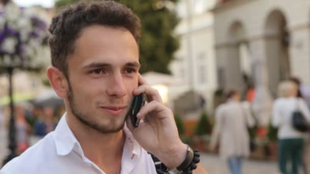 Улыбающийся 20-летний мужчина говорит о мобильнике на улице. Красивый парень говорит по телефону и делает красивую улыбку
. - Кадры, видео