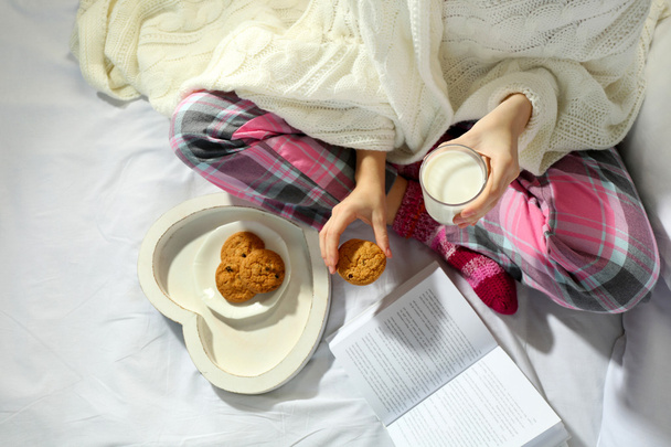Kobieta w piżamie czytając książkę i picie mleka z plików cookie na jej łóżko - Zdjęcie, obraz