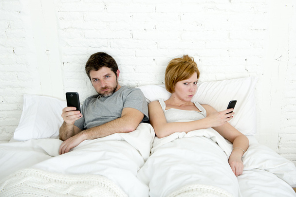 νεαρό παντρεμένο ζευγάρι χρησιμοποιώντας το κινητό τους τηλέφωνο στο κρεβάτι αγνοώντας μεταξύ τους σε προβλήματα σχέσεων επικοινωνίας - Φωτογραφία, εικόνα