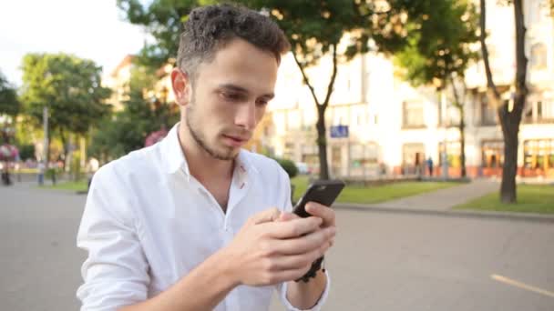 Young man in white shirt using smartphone outdoors. Guy texting at park. - Footage, Video
