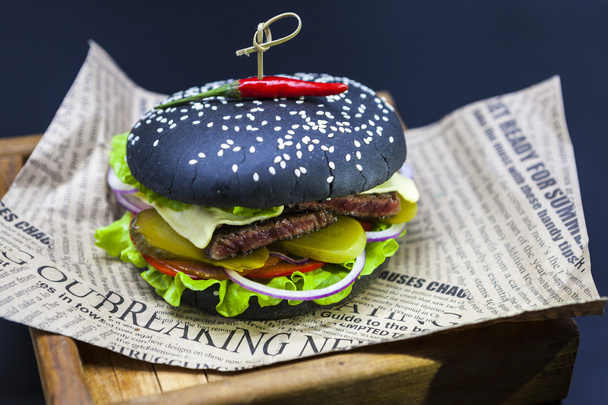 Black burger. A burger with a black roll slices of juicy marble beef, fused cheese, fresh salad and sauce of a barbecue. A burger on the newspaper on a wooden tray  on a dark background. - Fotó, kép