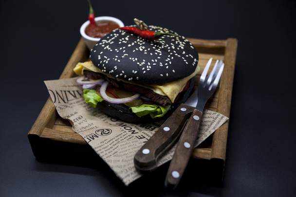 Black burger. A burger with a black roll slices of juicy marble beef, fused cheese, fresh salad and sauce of a barbecue. A burger on the newspaper on a wooden tray with a knife and a fork on a dark ba - Foto, afbeelding