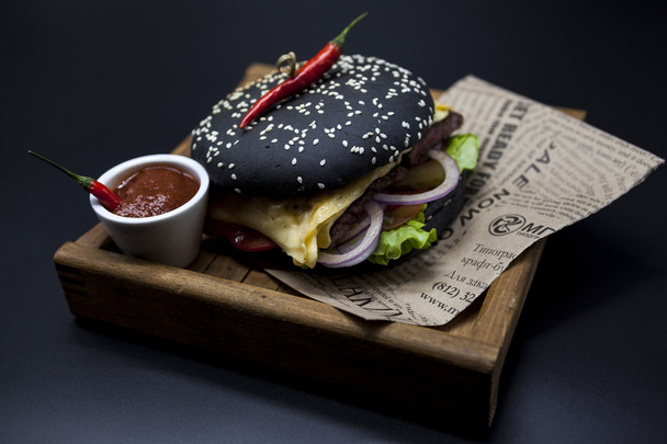 Black burger. A burger with a black roll slices of juicy marble beef, fused cheese, fresh salad and sauce of a barbecue. A burger on the newspaper on a wooden tray with a knife and a fork on a dark ba - Fotoğraf, Görsel