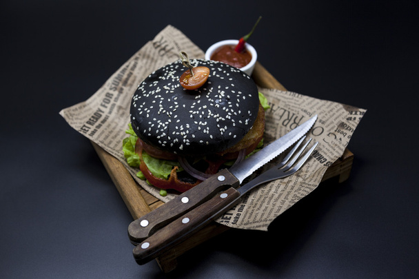 Black burger. A burger with a black roll slices of juicy marble beef, fused cheese, fresh salad and sauce of a barbecue. A burger on the newspaper on a wooden tray with a knife and a fork on a dark ba - Foto, Imagem
